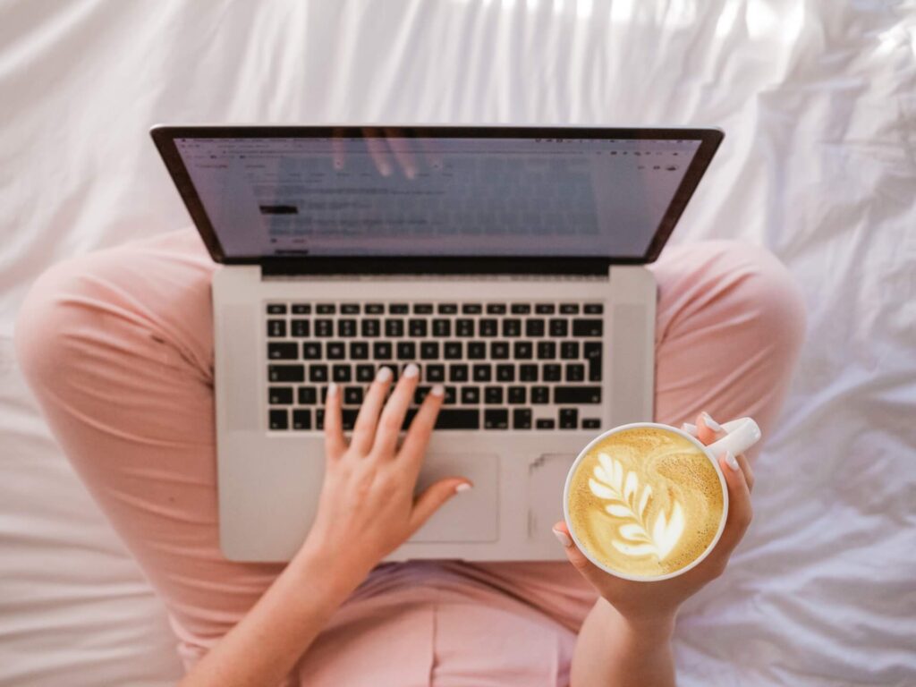 Woman typing on the laptop in bed and holding a cup of coffee