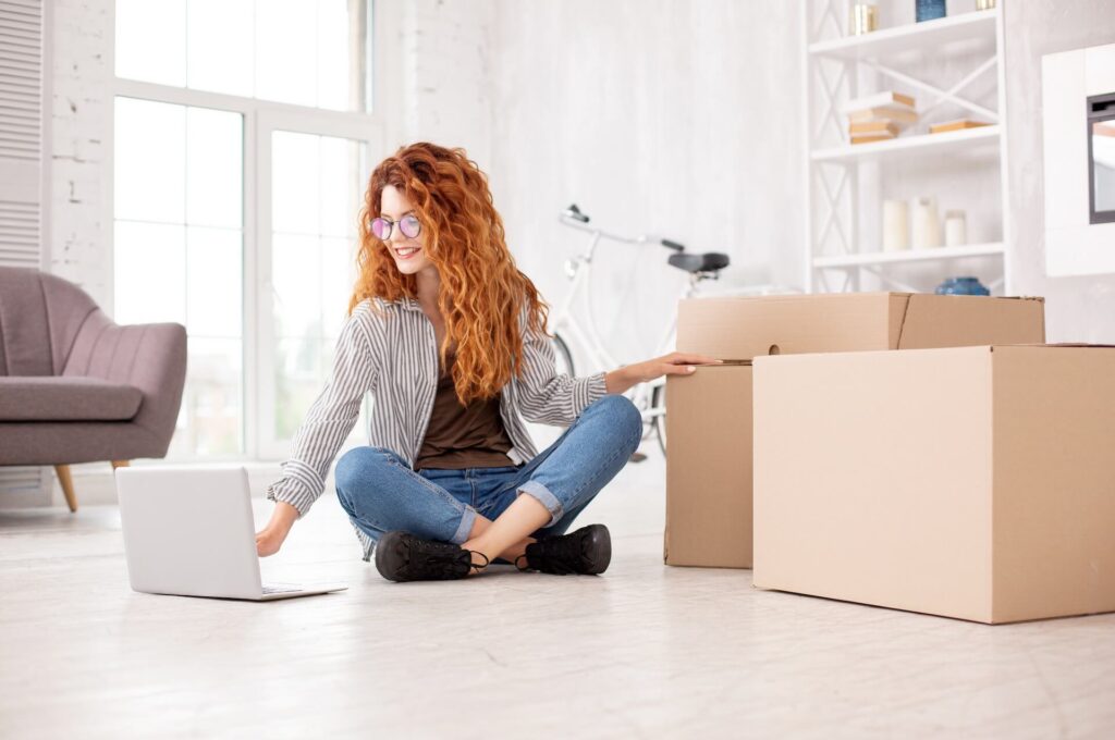 Woman sitting on the floor, looking at the laptop and boxes for moving overseas next to her