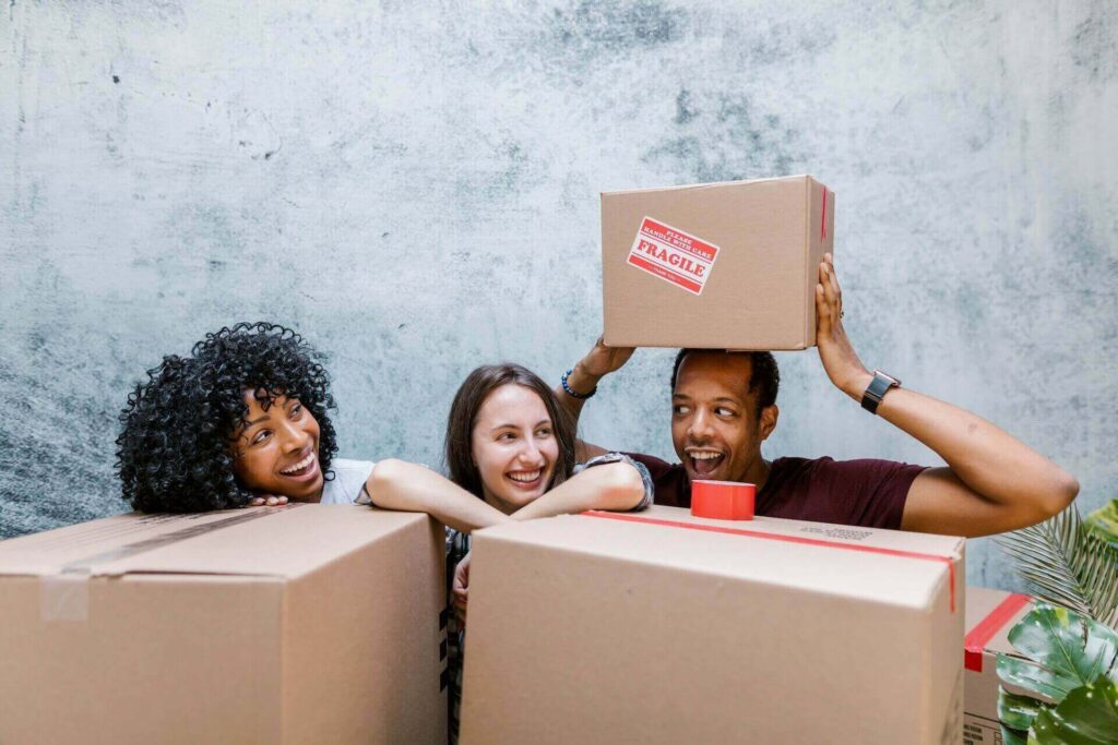 Three friends laughing behind a pile of boxes before moving internationally